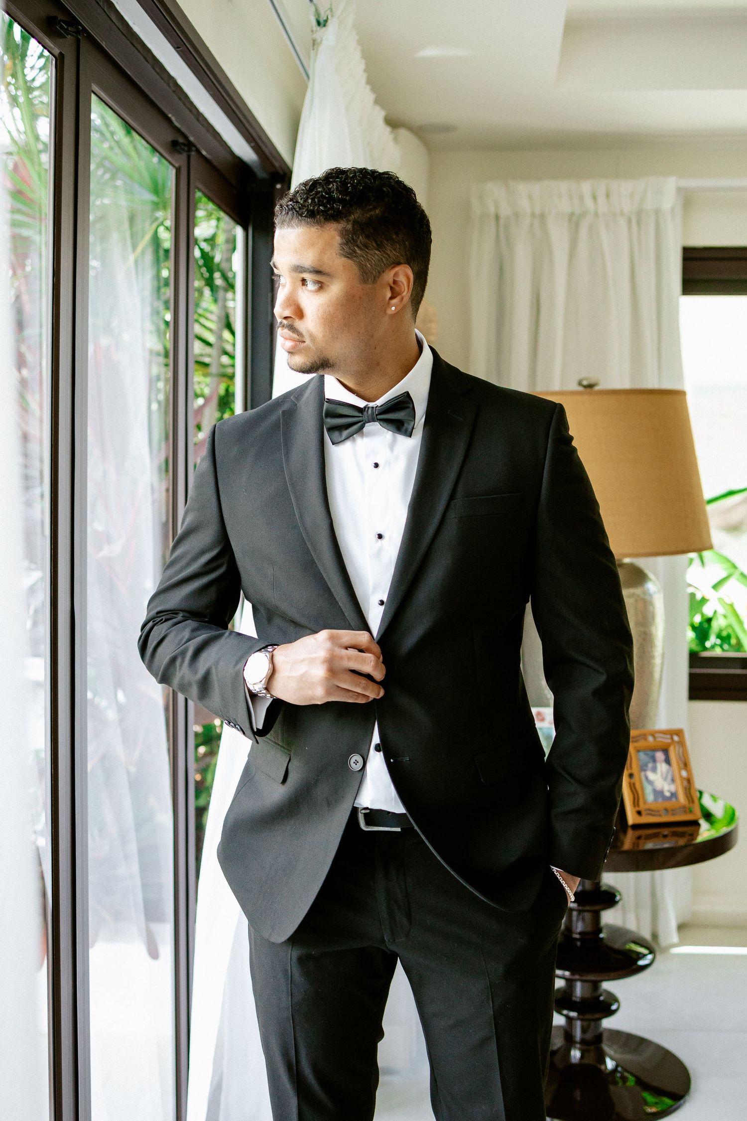 Handsome groom in black tuxedo and bow tie posing by glass doors on his wedding day.