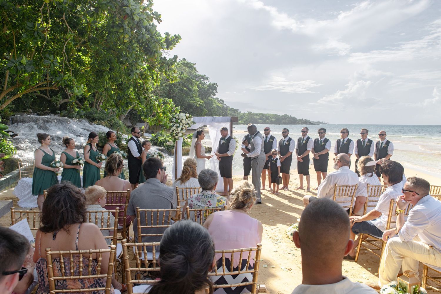 Beach wedding ceremony with bridal party and guests seated on sandy shore by the ocean.