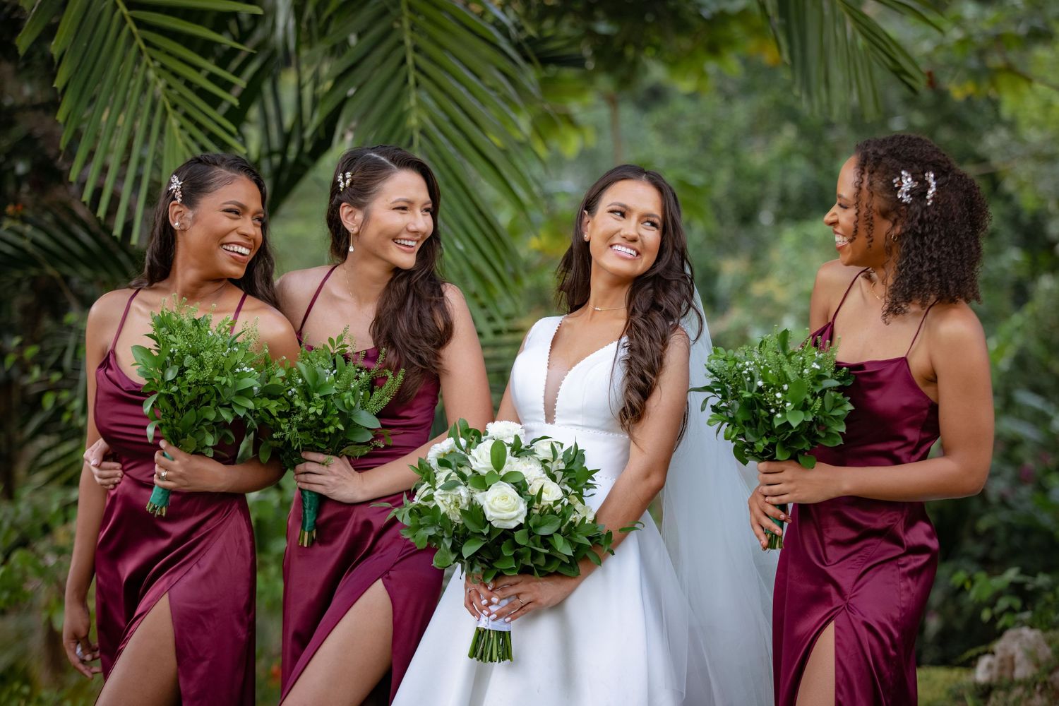 Bride in white gown with three bridesmaids in burgundy dresses holding green bouquets outdoors.