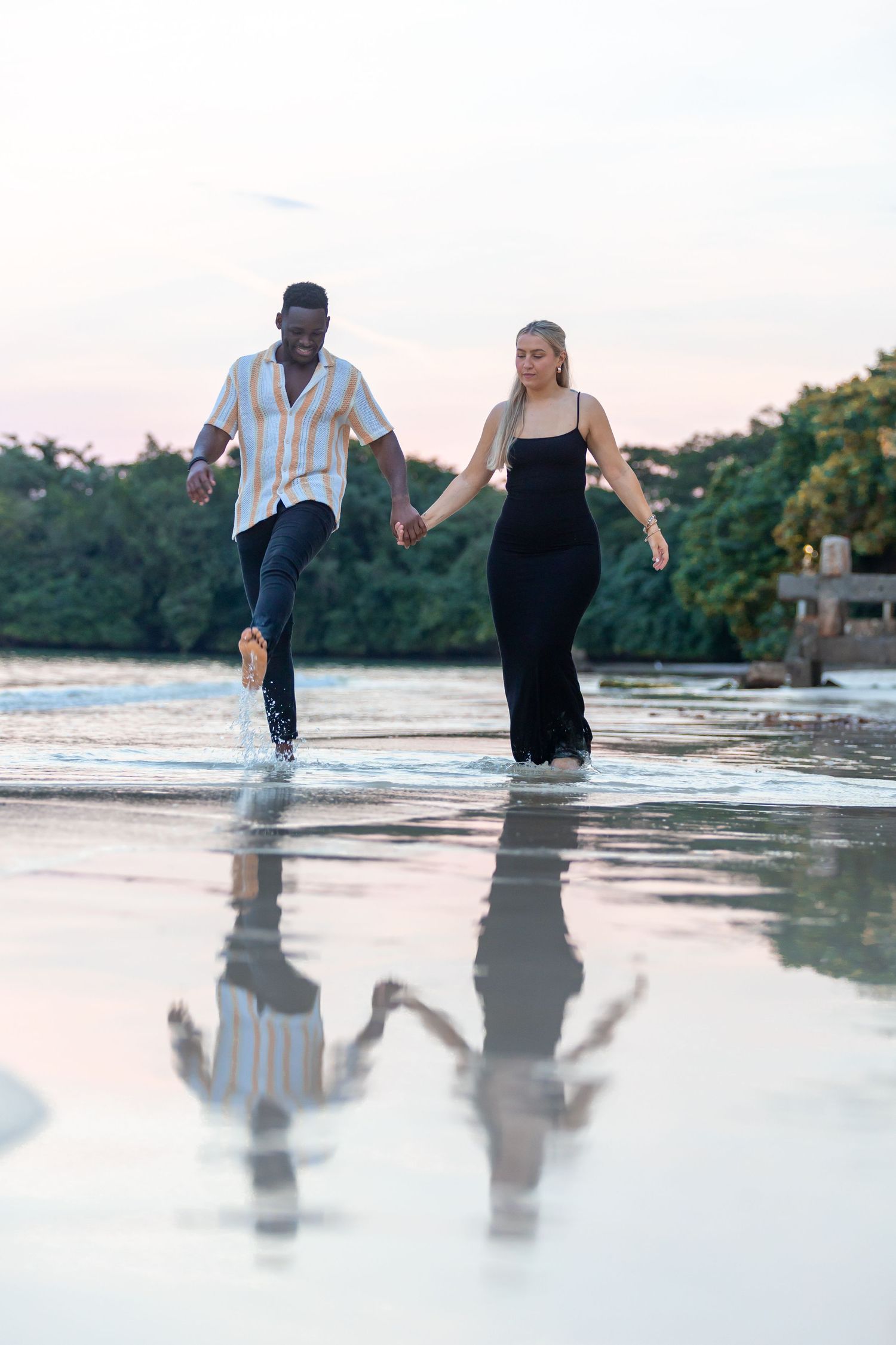 Joyful interracial couple running hand in hand through shallow beach water at sunset.