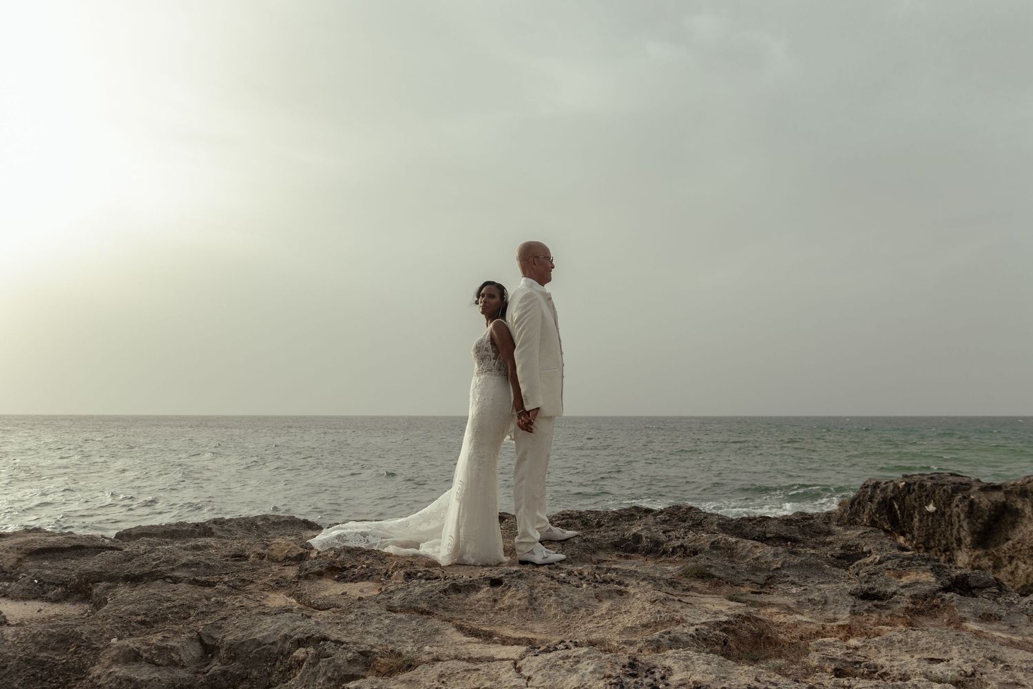 Bride and groom in white wedding attire stand on rocky oceanside cliff at sunset.
