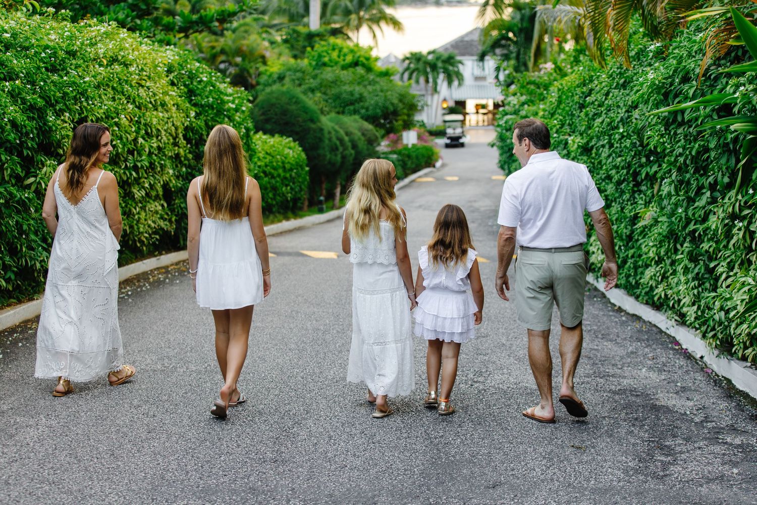Family of five in white outfits walking down a tropical resort path surrounded by lush green foliage.