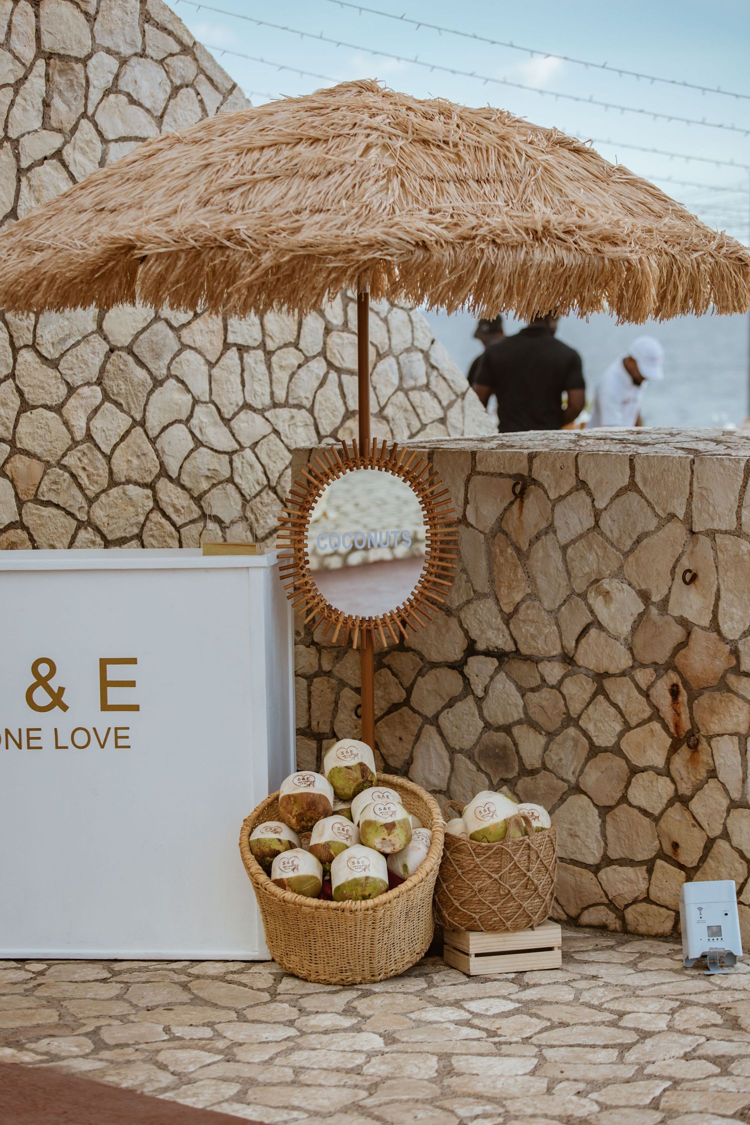 Boho beach wedding setup with rattan umbrella, wicker baskets, monogram sign, and stone wall backdrop.
