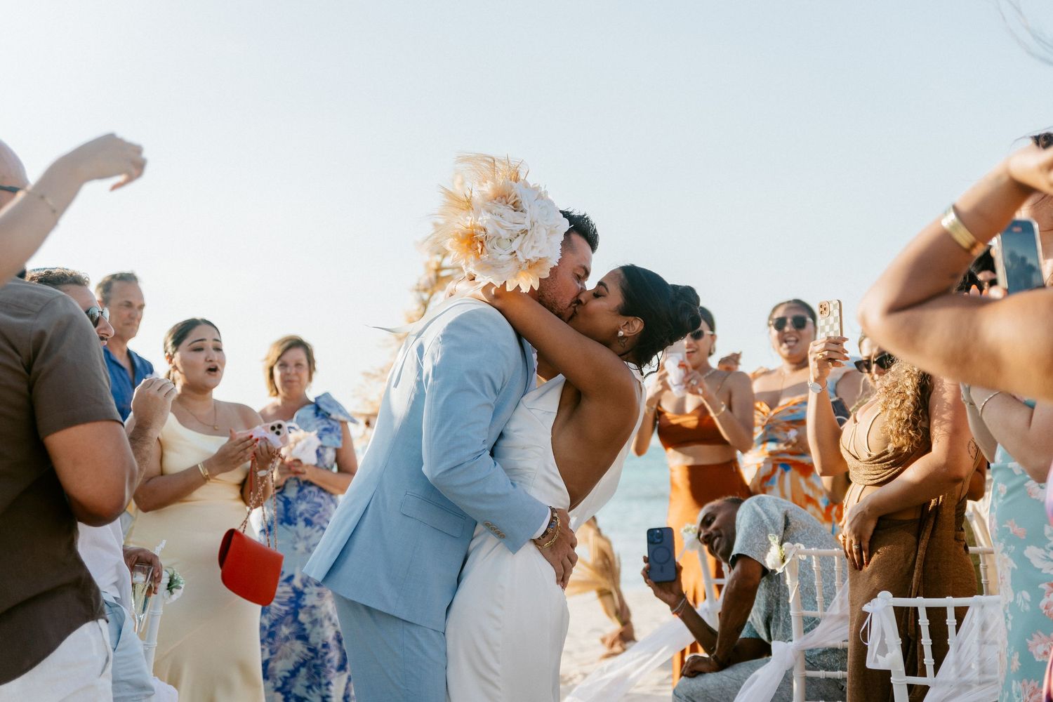 Couple shares a romantic first kiss at outdoor beach wedding ceremony surrounded by cheering guests in summer attire.