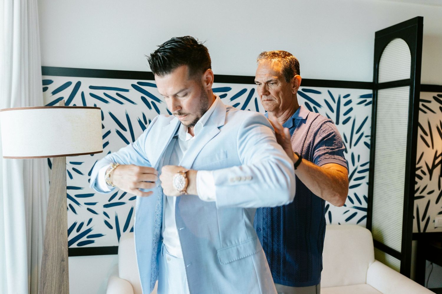 Groom in light blue suit adjusting cufflinks while older man helps with jacket in elegant room.