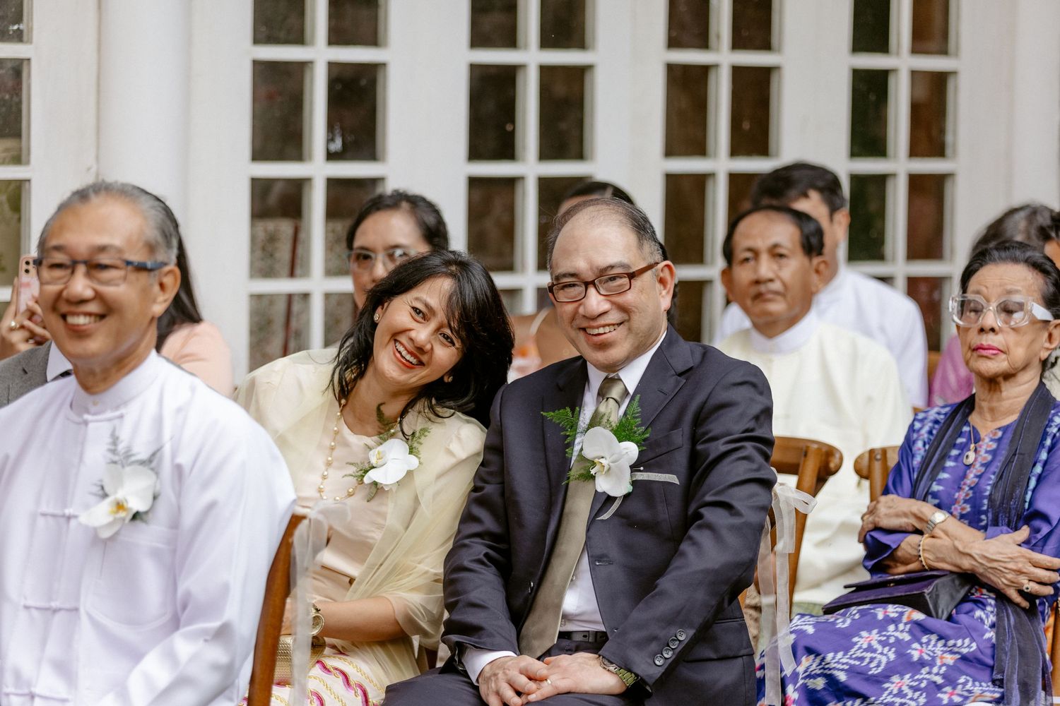 Smiling wedding guests seated outdoors, including a couple in formal attire with boutonnieres, at an elegant garden ceremony.