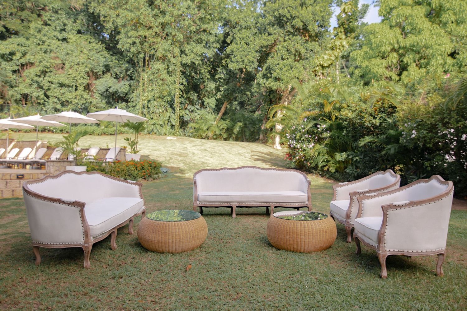 Elegant outdoor lounge area with white upholstered sofas and wicker coffee tables on a green lawn surrounded by lush trees.