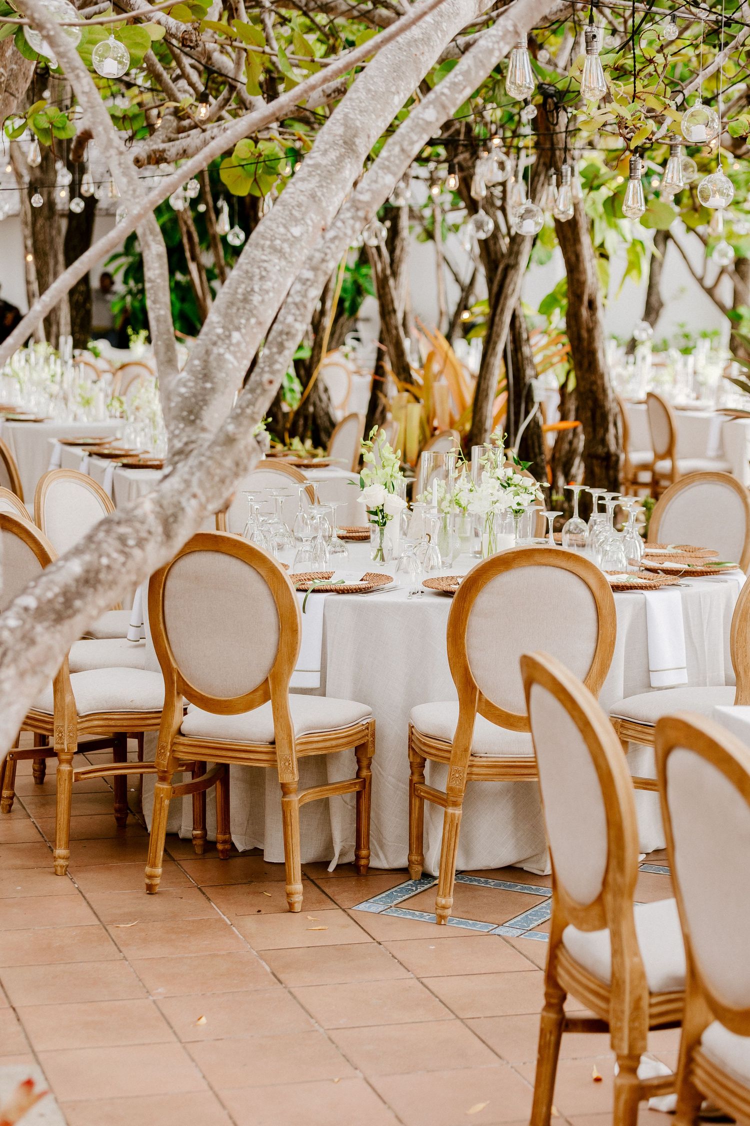 Elegant outdoor wedding reception with gold Louis chairs, white linen tables, and hanging glass orbs under lush tropical trees.