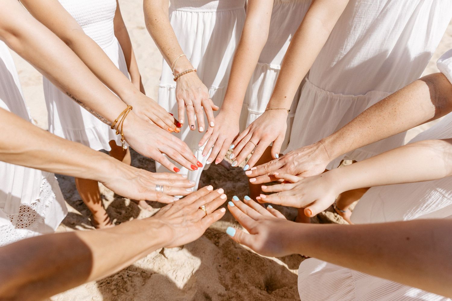 Diverse group of women in white dresses forming a circle with hands together, showcasing unity and friendship.