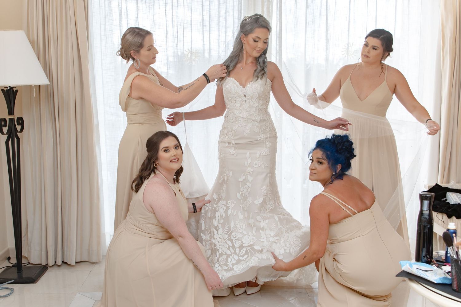 Bride in lace wedding gown surrounded by four bridesmaids in champagne dresses getting ready.