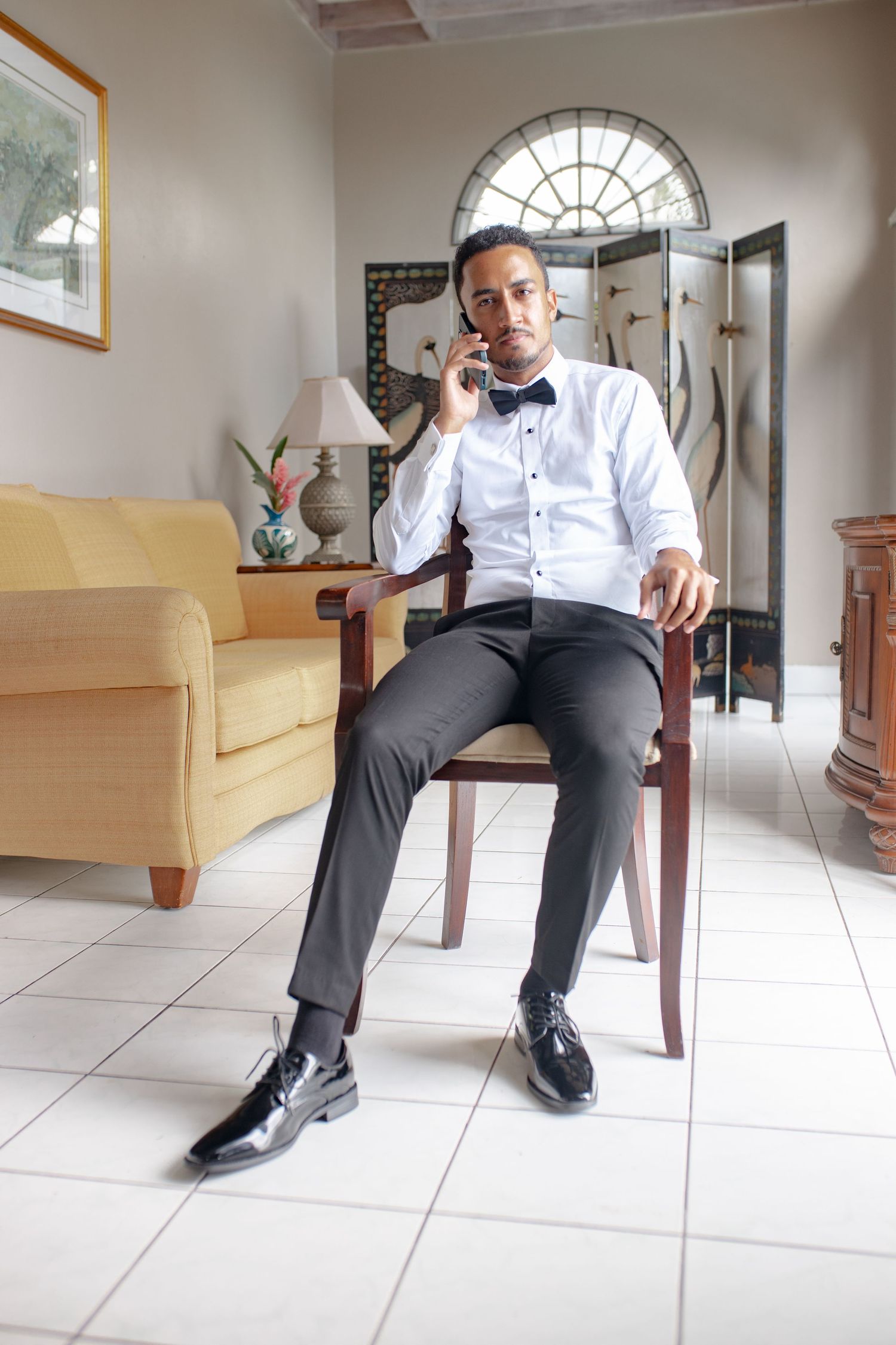 Groom in white dress shirt and bow tie sitting in chair talking on smartphone indoors.