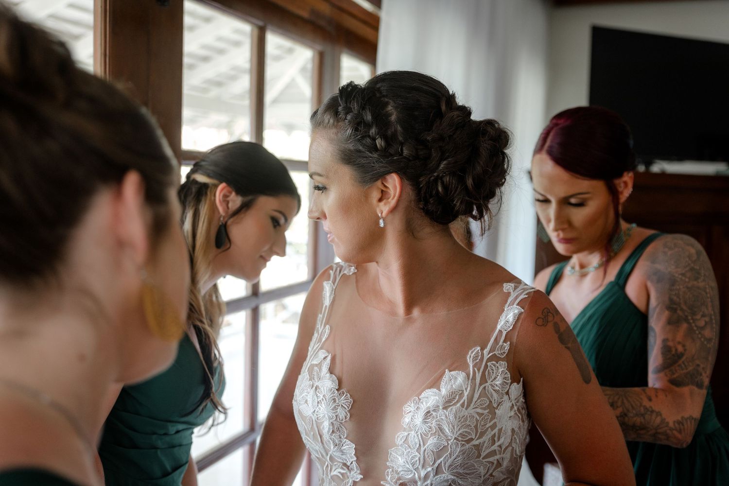 Bridesmaids in emerald green dresses help bride in lace wedding gown prepare for her ceremony.