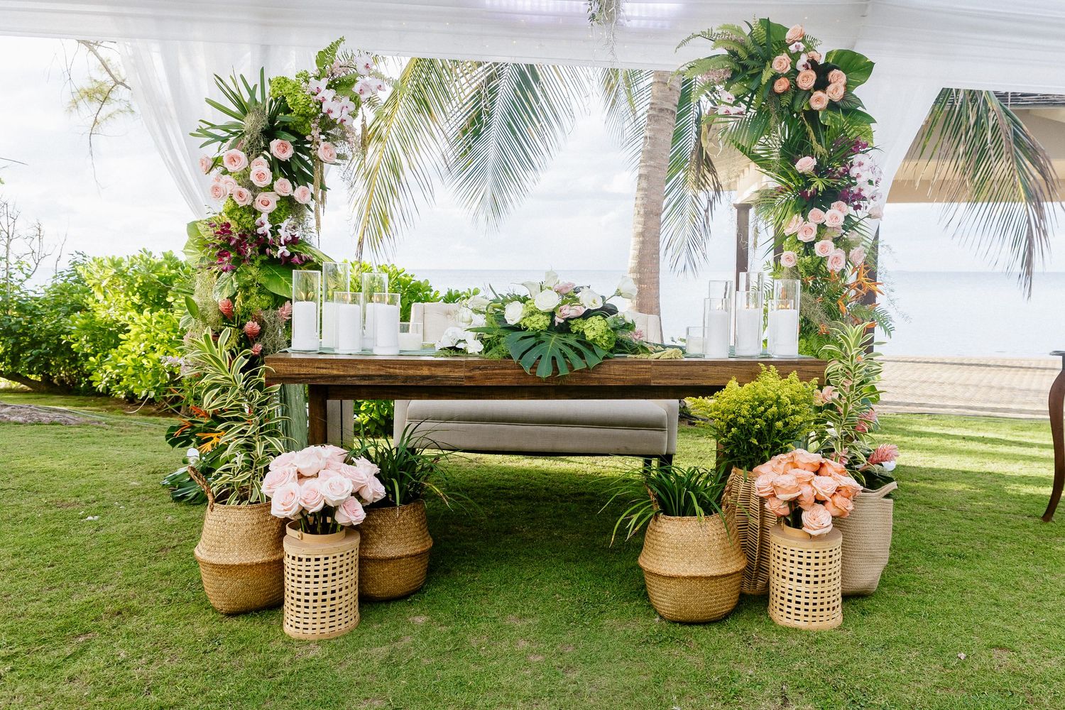 Tropical wedding welcome table with floral arch, candles, and wicker baskets on green lawn near palm trees.