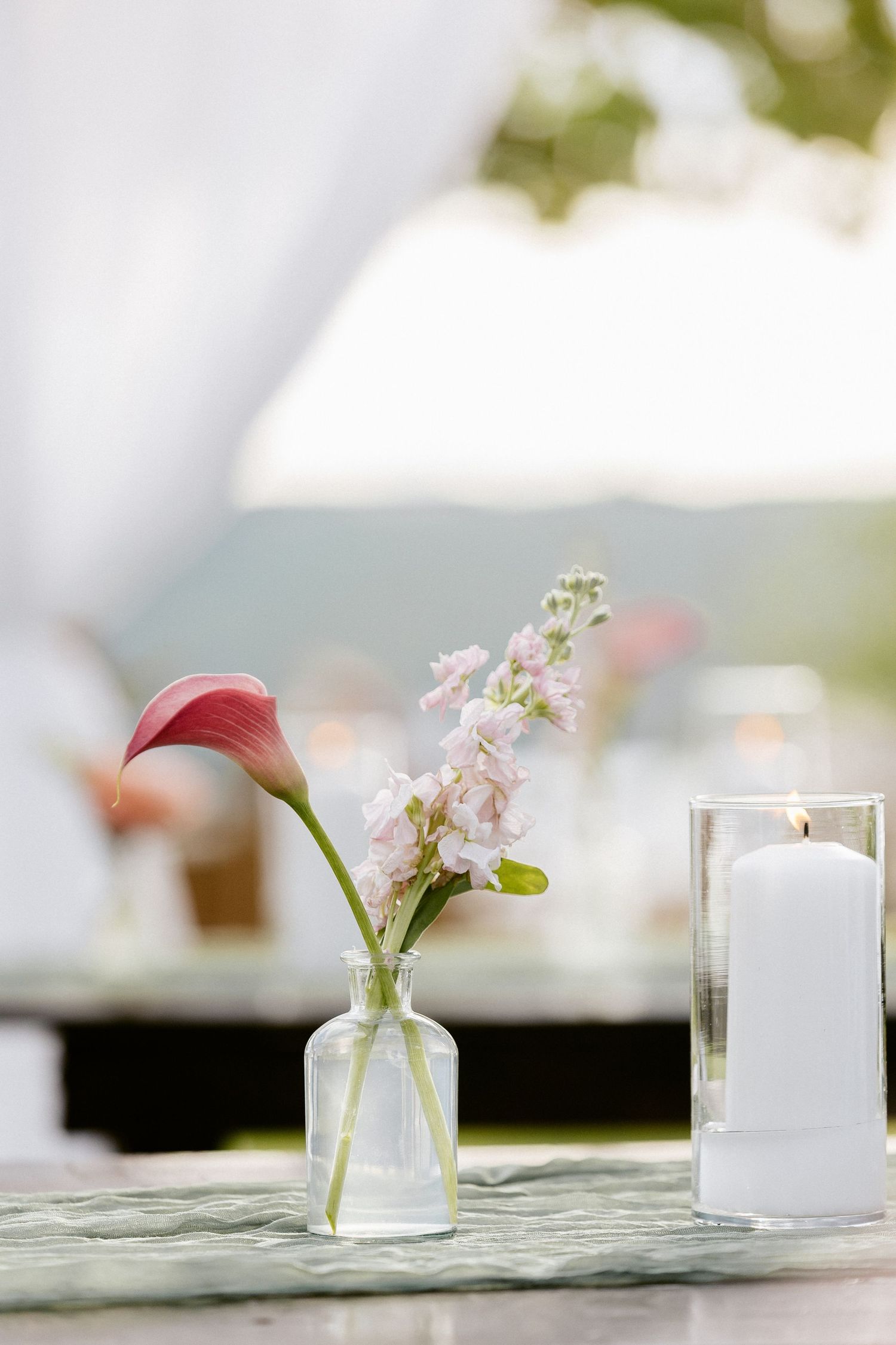 Elegant wedding table with a pink calla lily in a glass vase, small white flowers, and a lit pillar candle outdoors.