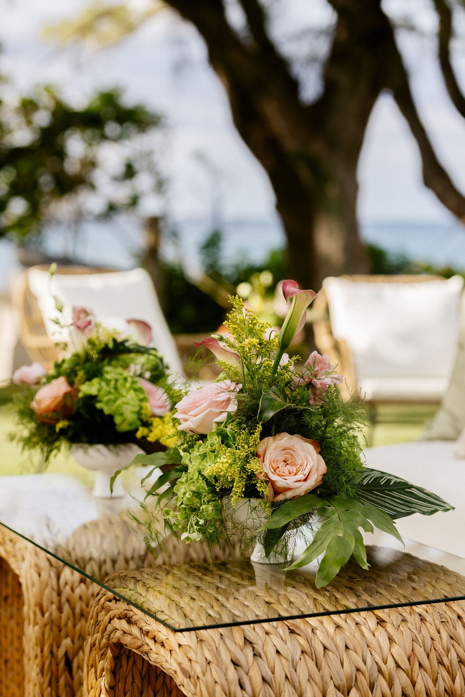 Elegant floral arrangements with pink roses and greenery in white vases on a wicker glass-top table at an outdoor event.