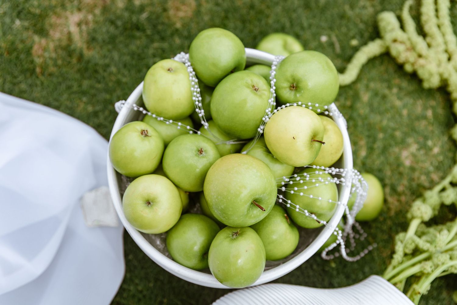A white bowl filled with fresh green apples decorated with pearl bead garlands, placed on green grass.