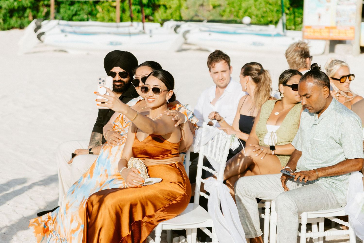 Wedding guests in sunglasses take selfies while seated at an outdoor beach ceremony on white chairs.