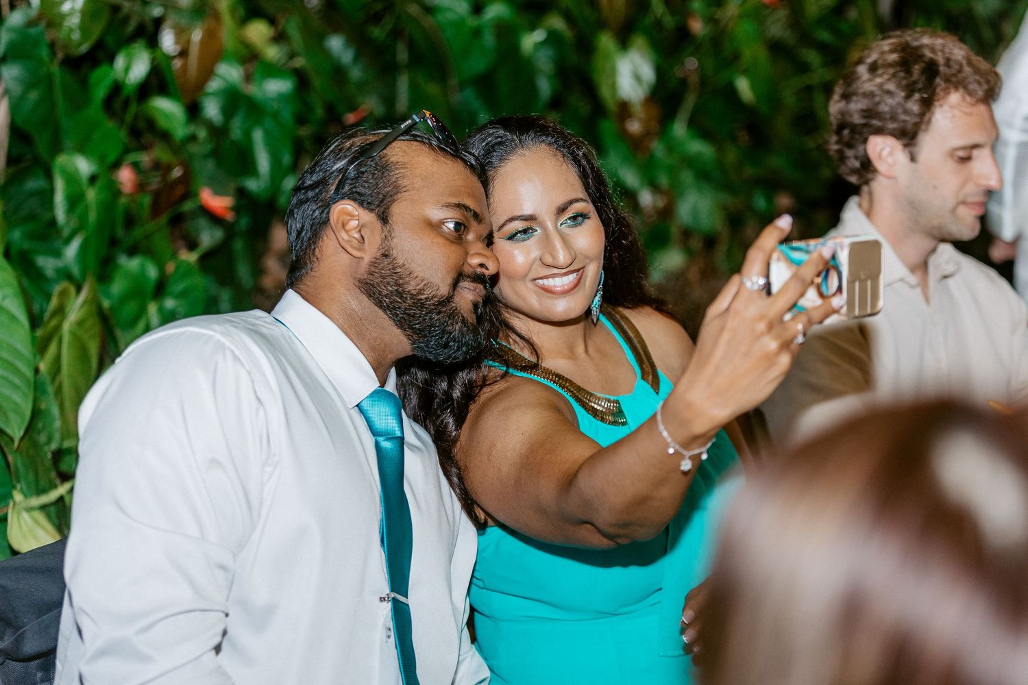 Man and woman taking a selfie at a wedding reception surrounded by lush green foliage.