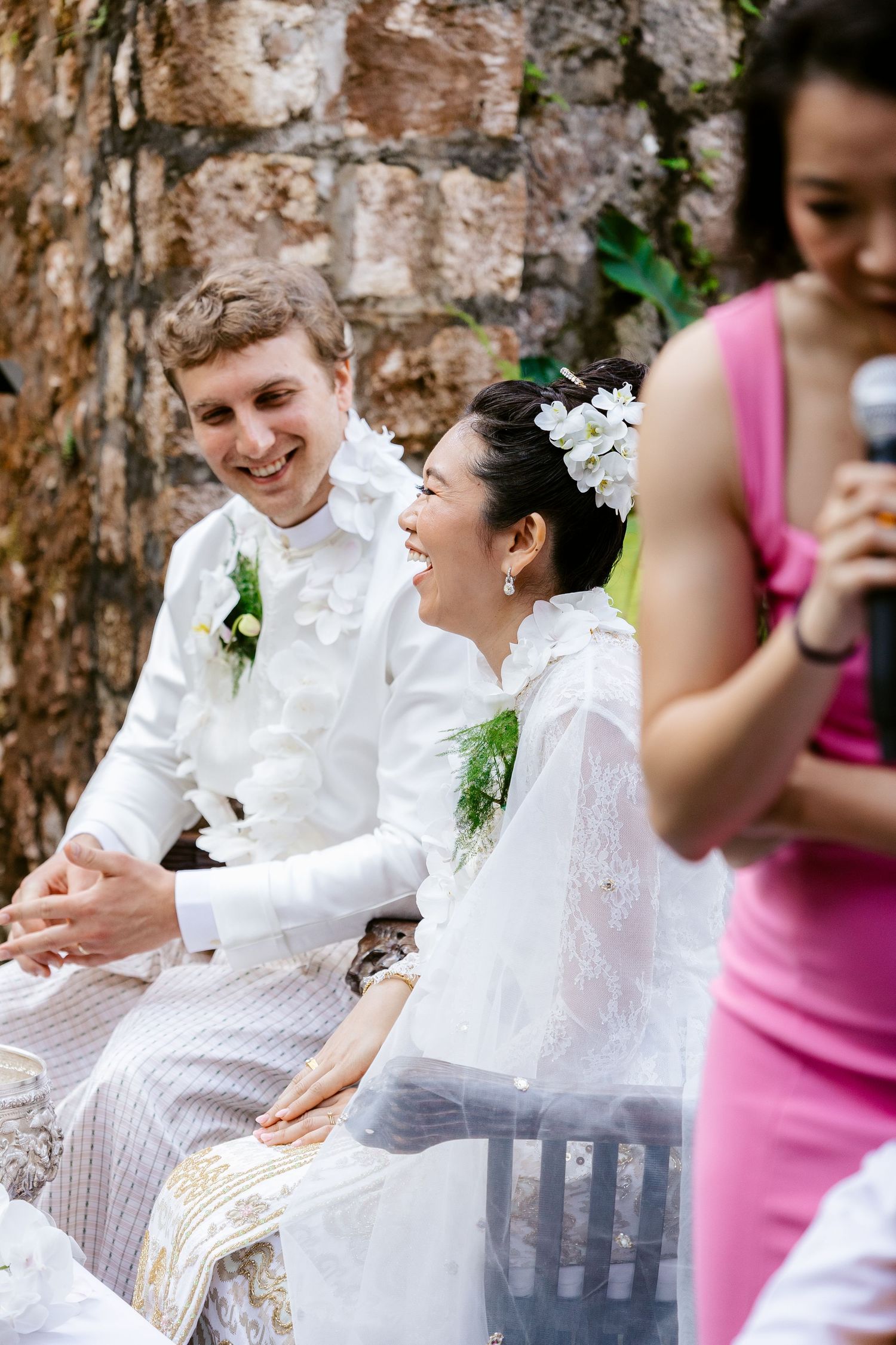 Smiling bride and groom in white traditional wedding attire laughing during outdoor ceremony.
