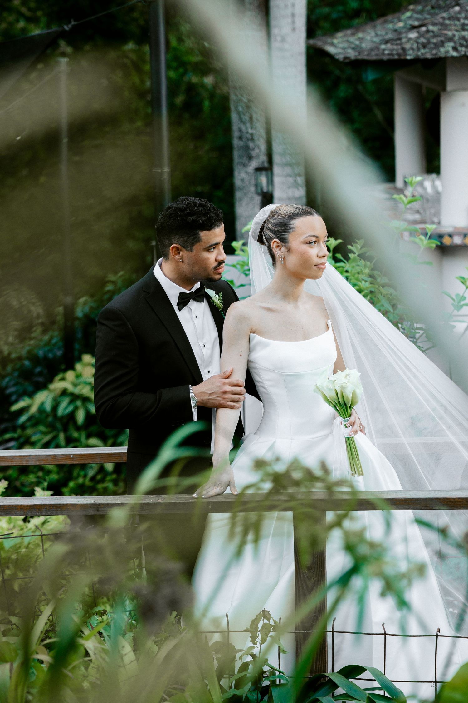 A bride in a white gown and veil holds a bouquet while a groom in a black tuxedo stands beside her outdoors.