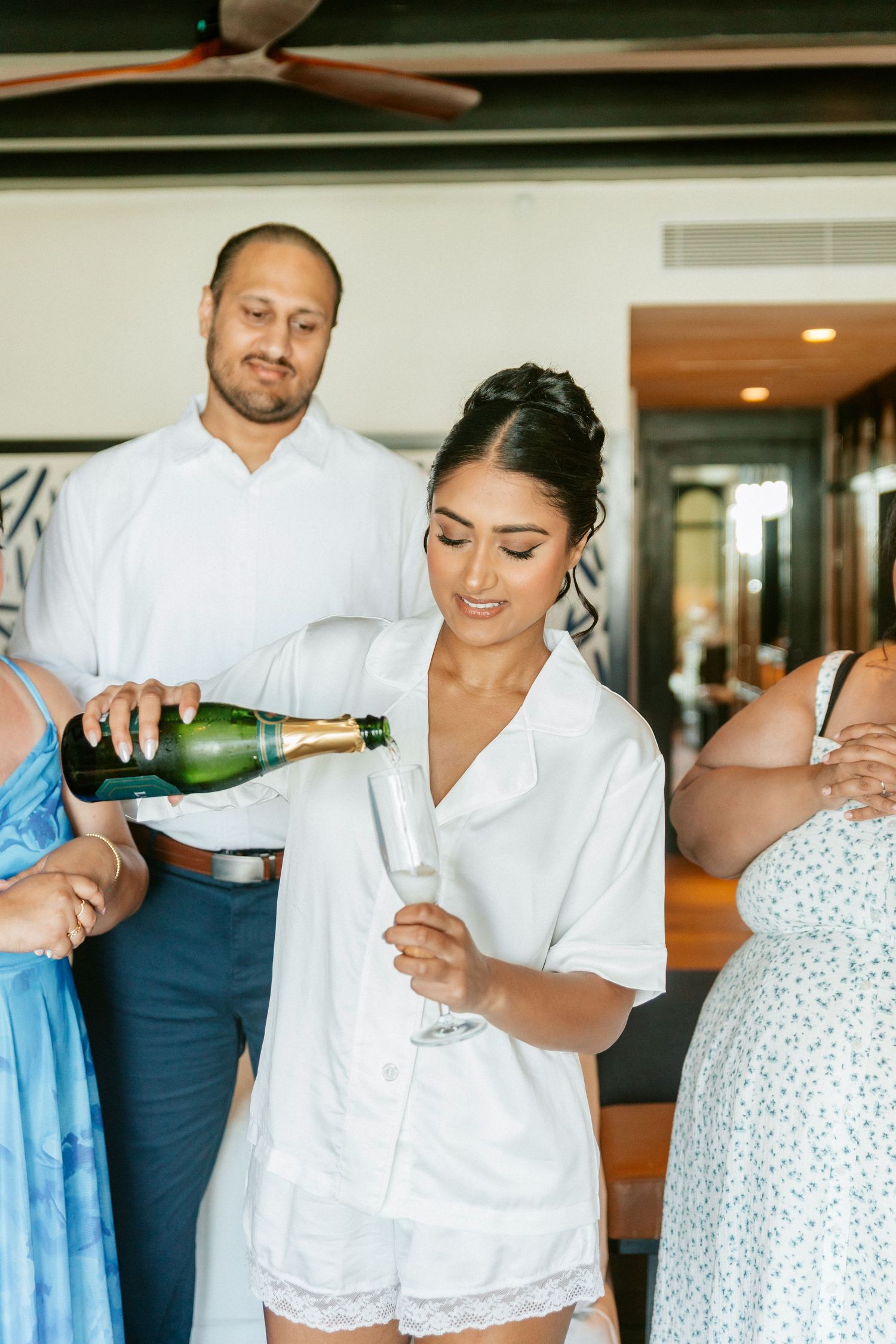 Bride in white pajamas receiving champagne pour during wedding morning preparations with guests nearby.