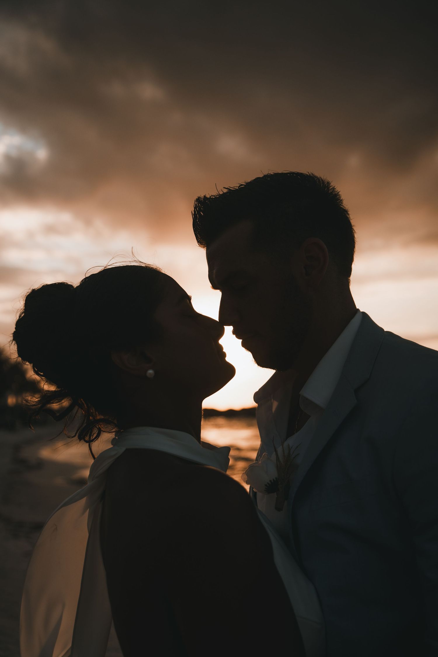 Romantic silhouette of bride and groom about to kiss against dramatic sunset sky.