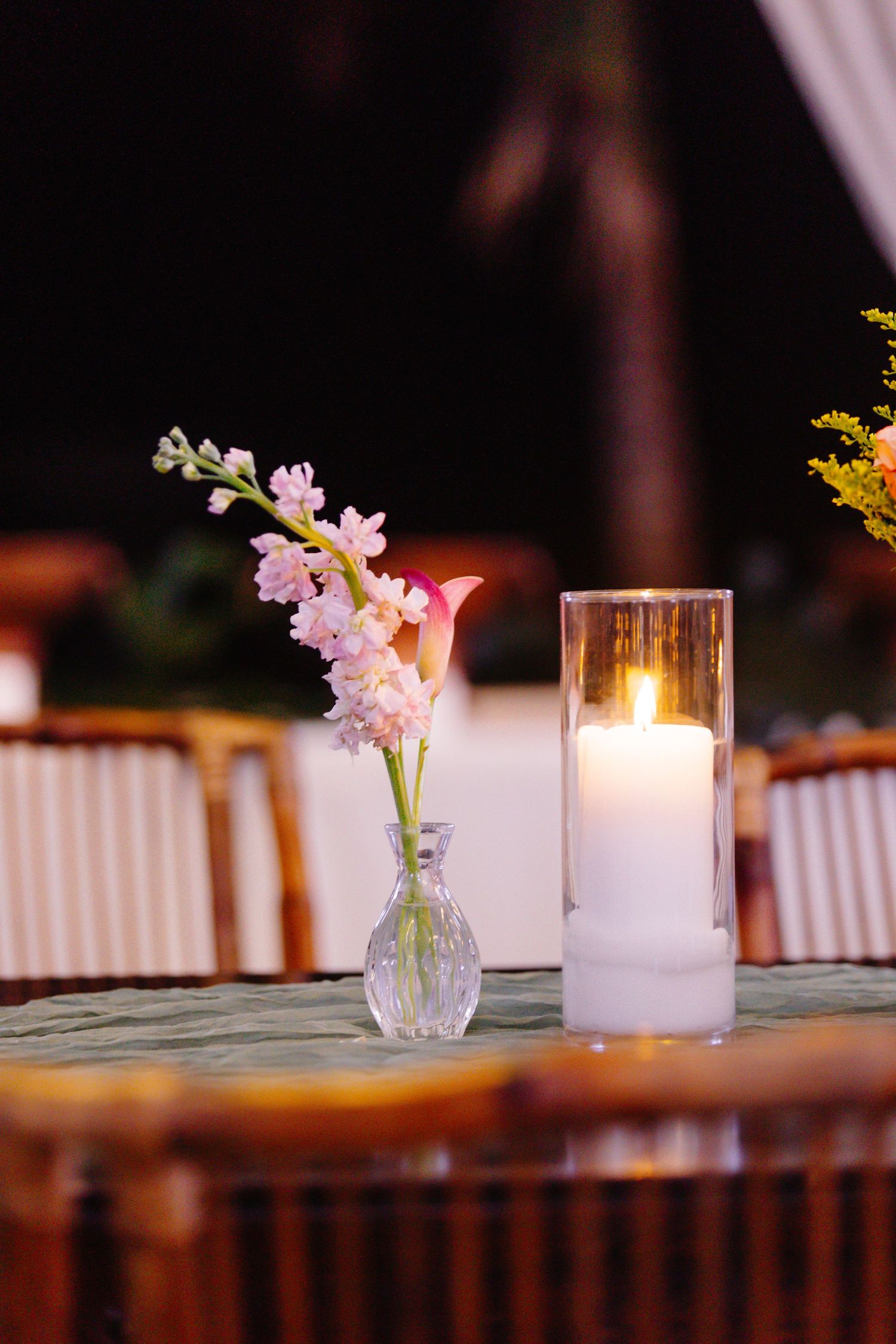 Glass bud vase with pink flowers beside a lit white pillar candle in a glass holder on a wedding reception table.
