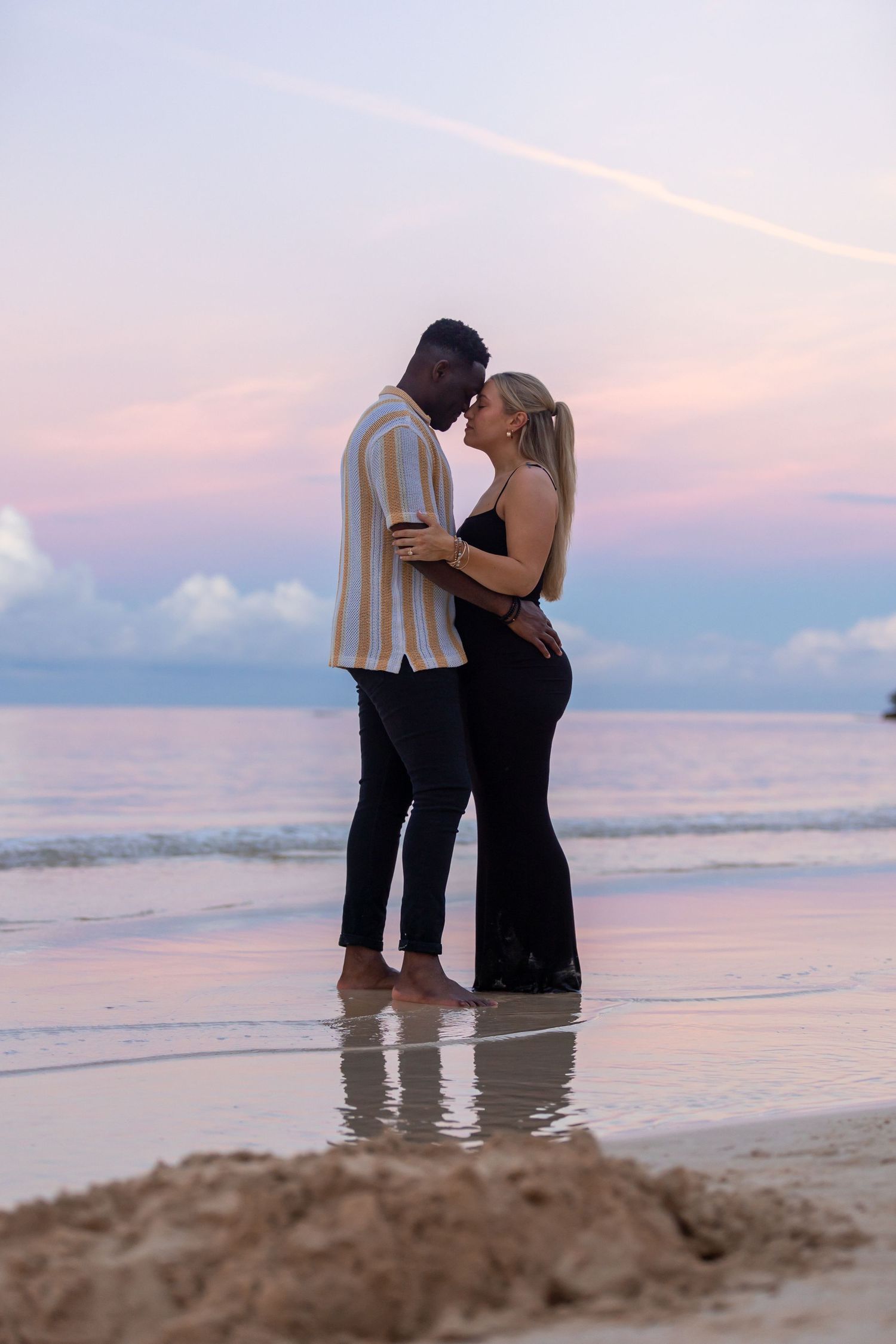 Romantic interracial couple standing close together on a beach during a pink and blue sunset.
