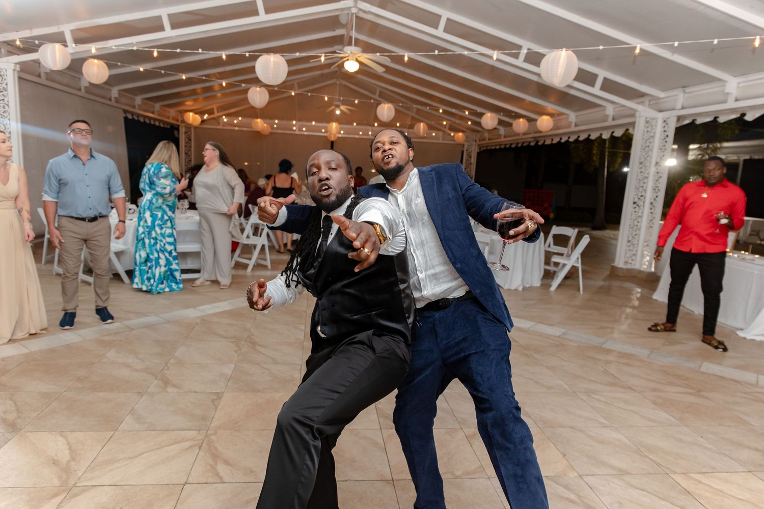 Two men in suits show off energetic dance moves at wedding reception under paper lantern lights.