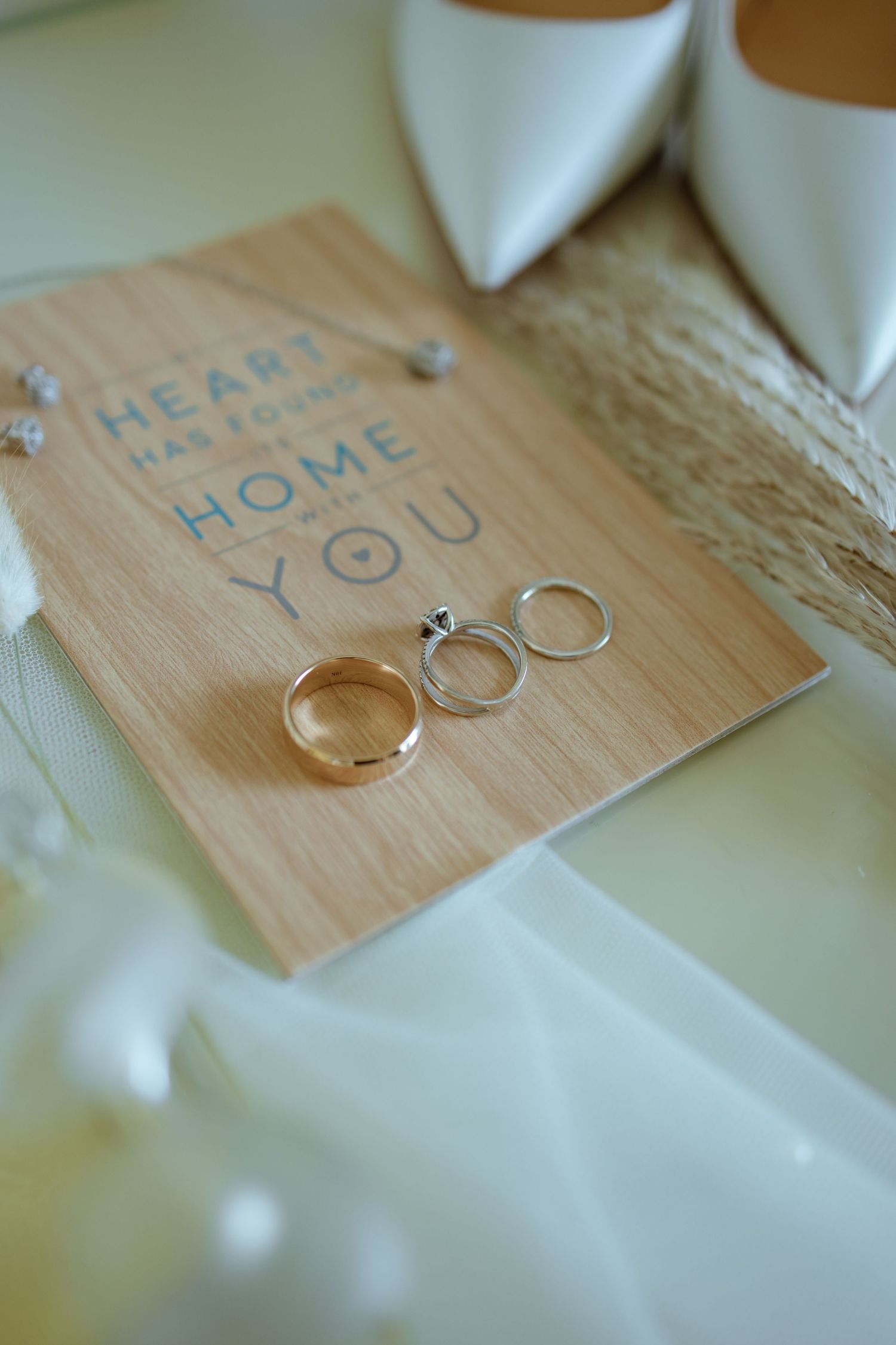 Wedding rings and engagement ring displayed on wooden card with white bridal heels and veil.