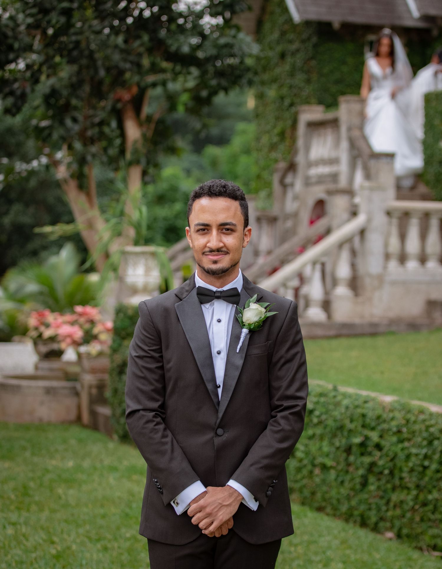 Smiling groom in black tuxedo with boutonniere as bride descends garden staircase behind him.