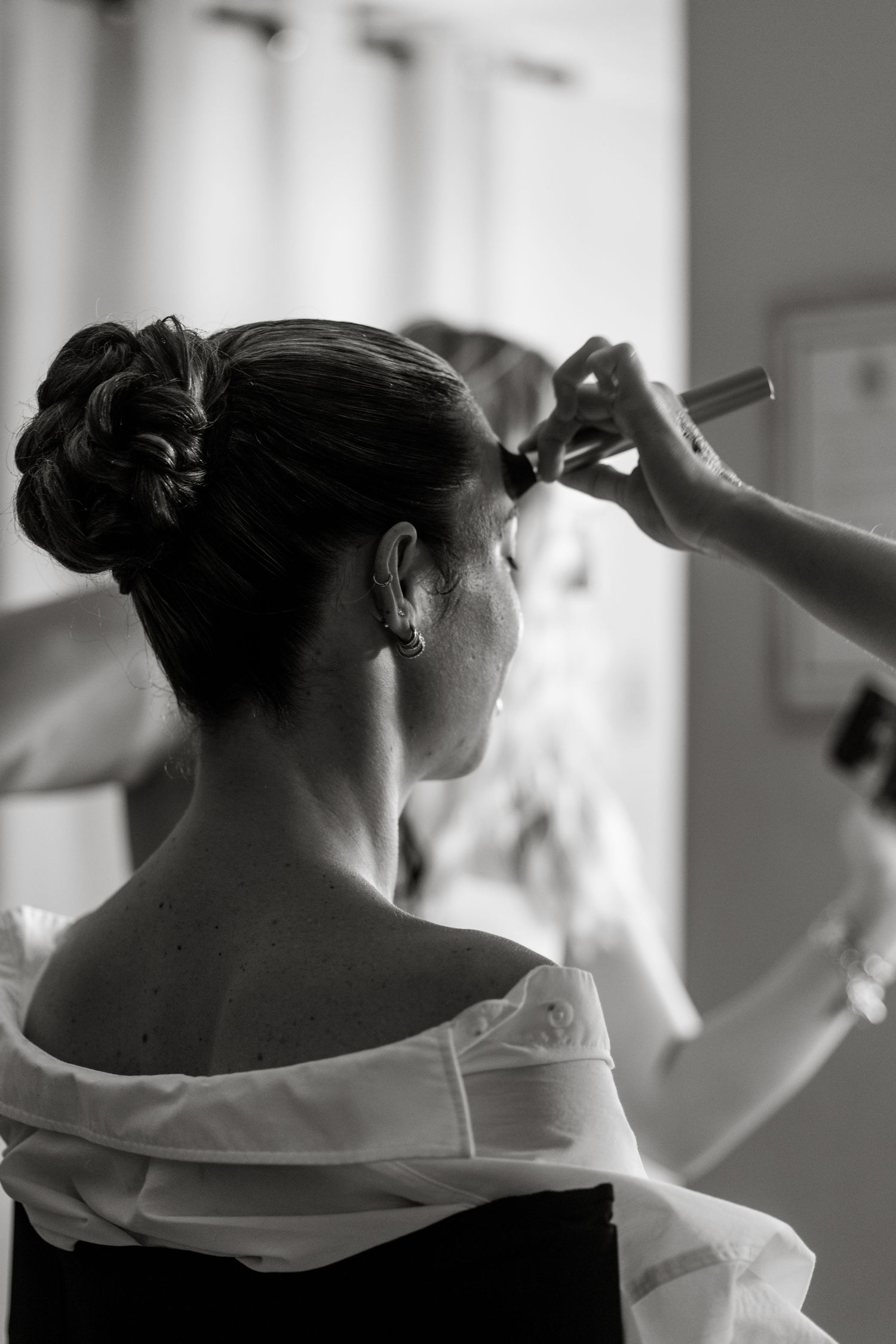 Black and white photo of bride getting makeup applied with hair in elegant updo before wedding.