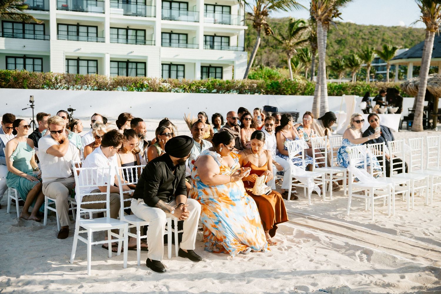 Guests seated in white chairs on a sandy beach wedding ceremony with a resort building and palm trees in the background.