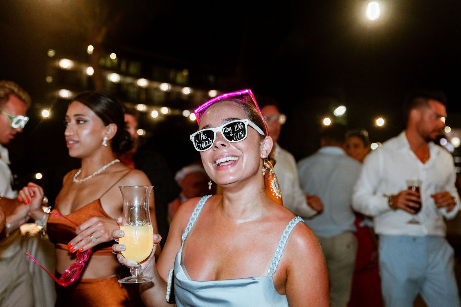 Wedding guest wearing pink party hat and white sunglasses smiling at outdoor evening reception.