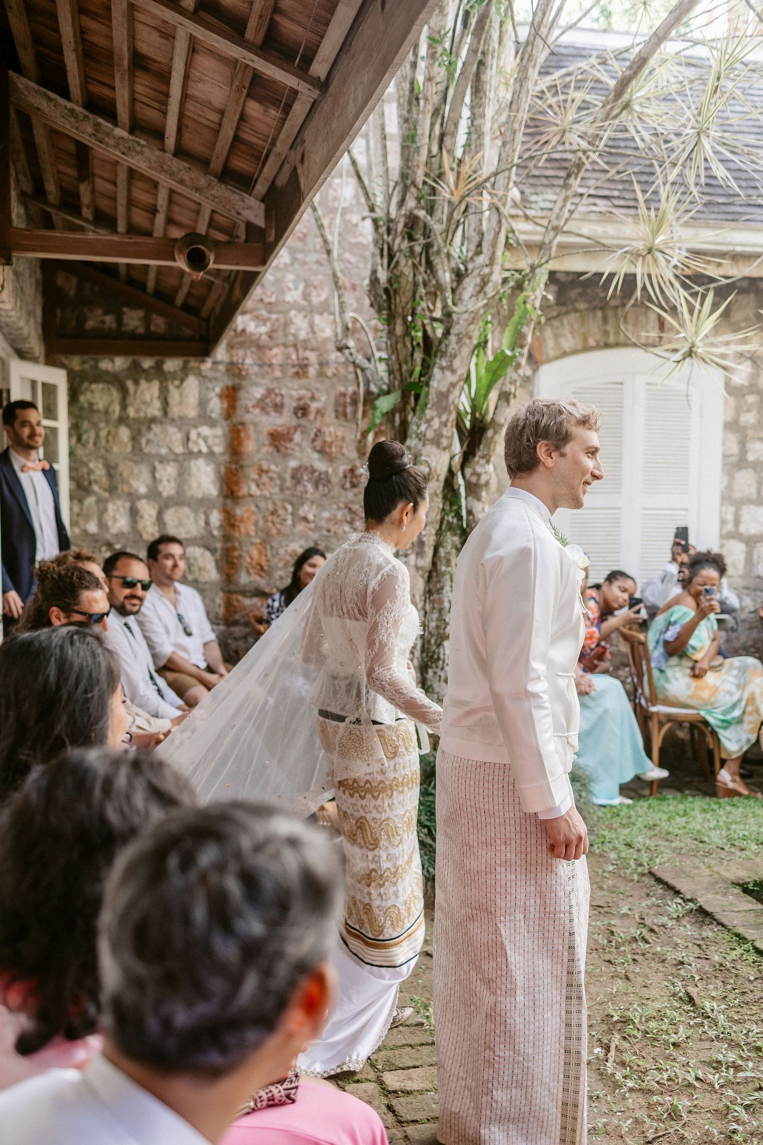 Bride and groom in traditional white attire walking during an outdoor wedding ceremony with seated guests.
