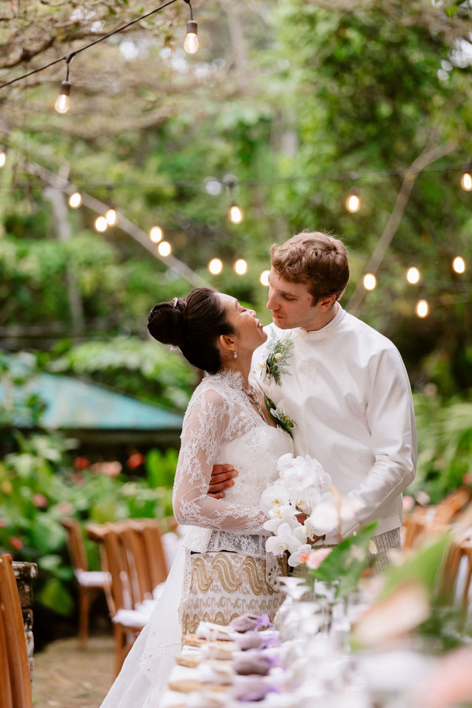 Bride and groom share romantic moment at outdoor wedding reception table adorned with string lights and florals.