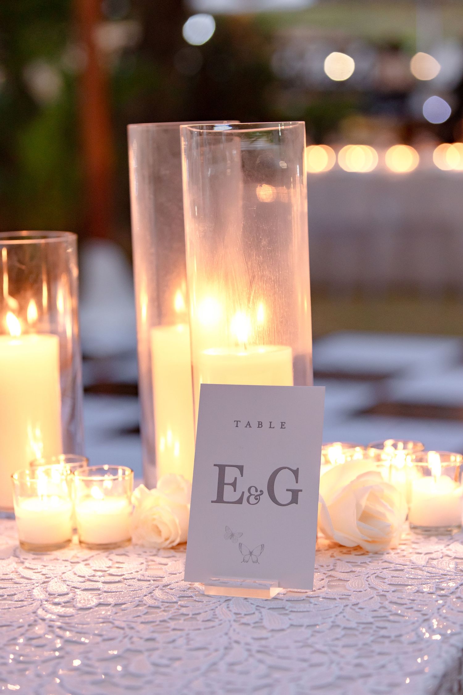 Wedding table card displaying initials E&G surrounded by glowing white candles and roses on a lace tablecloth.