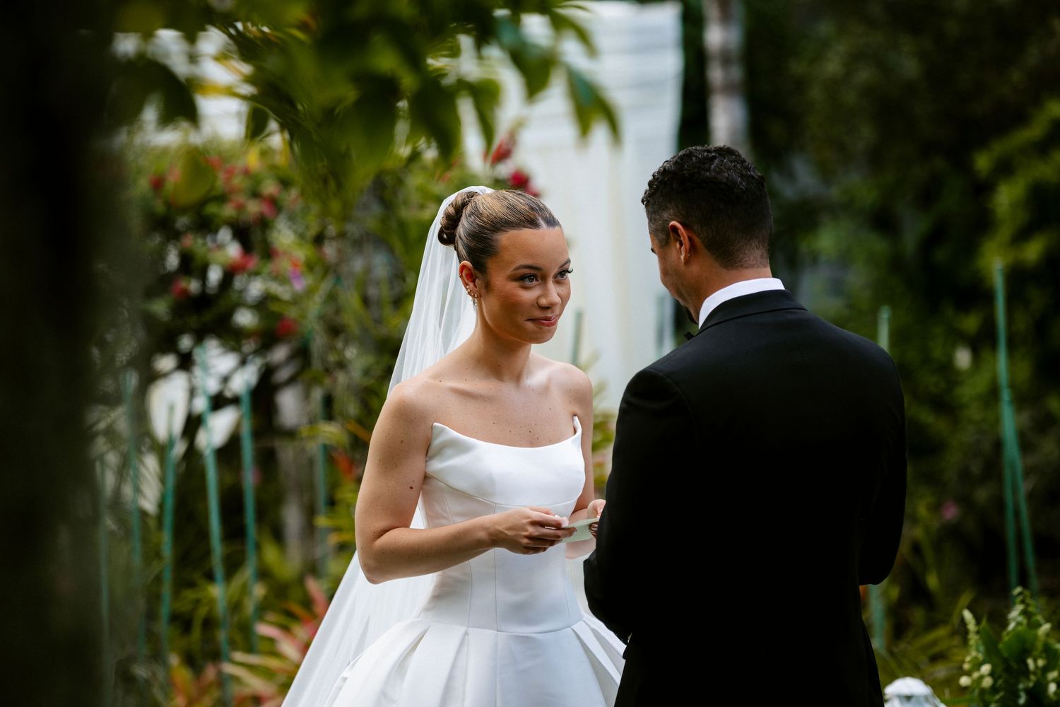 Bride in white strapless gown and veil exchanging vows with groom in black suit at outdoor garden wedding.