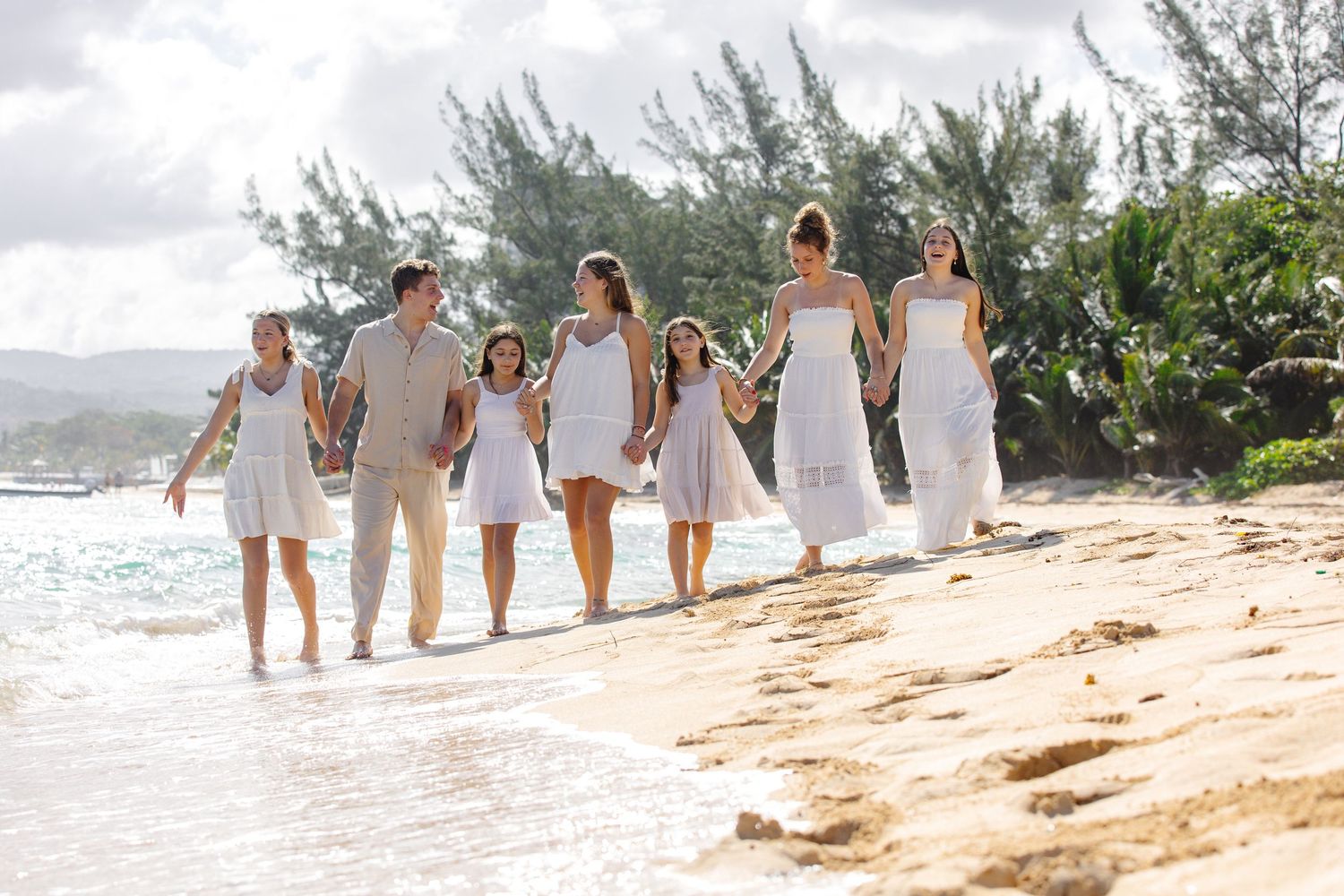 A joyful family in white outfits walks hand-in-hand along a tropical sandy beach with lush green trees.