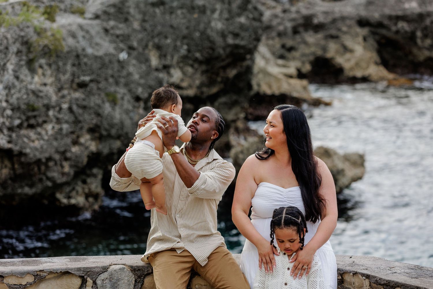 Family of four posing by rocky coastline, father lifting baby while mother holds toddler in white dress outdoors.