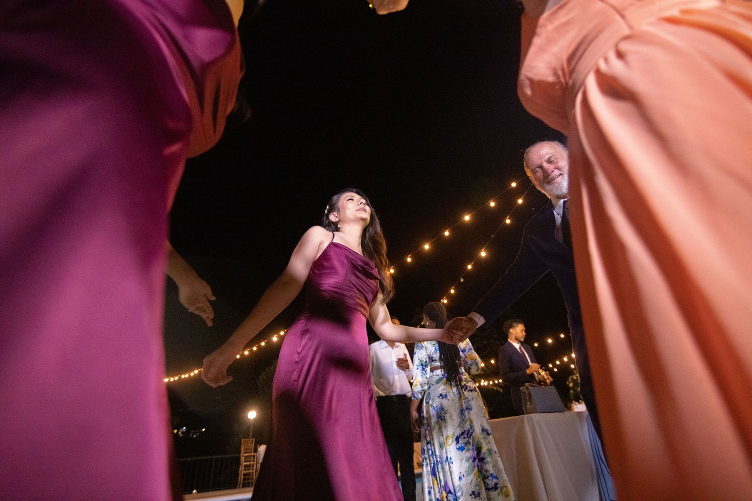 Wedding guests in purple and peach gowns dancing under string lights at night.