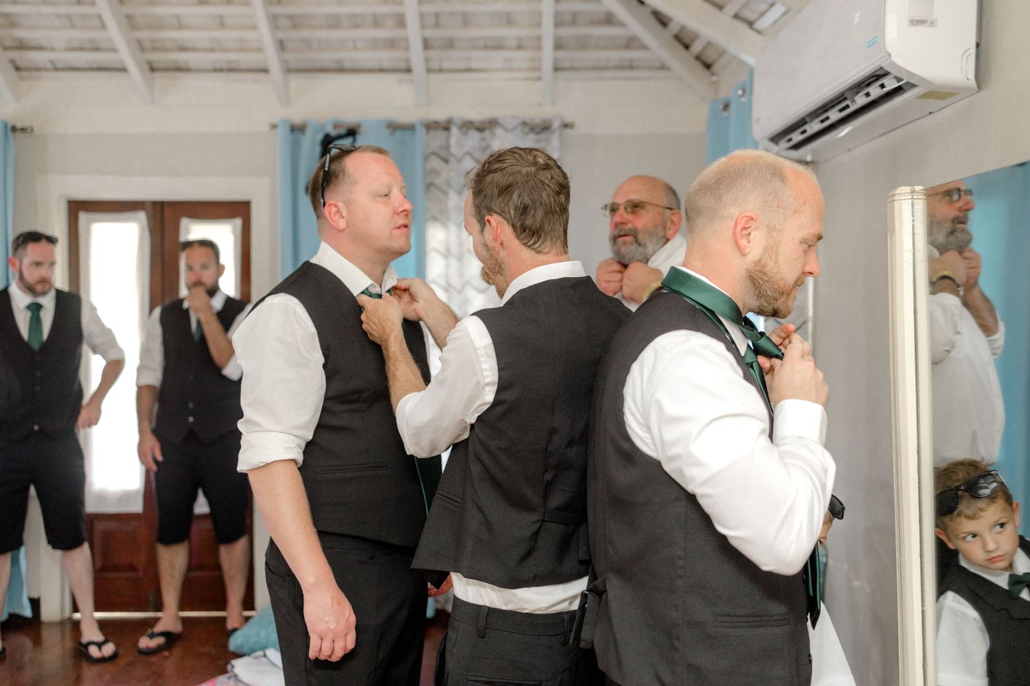 Groomsmen help each other adjust bow ties and neckties while getting ready for a beach wedding.