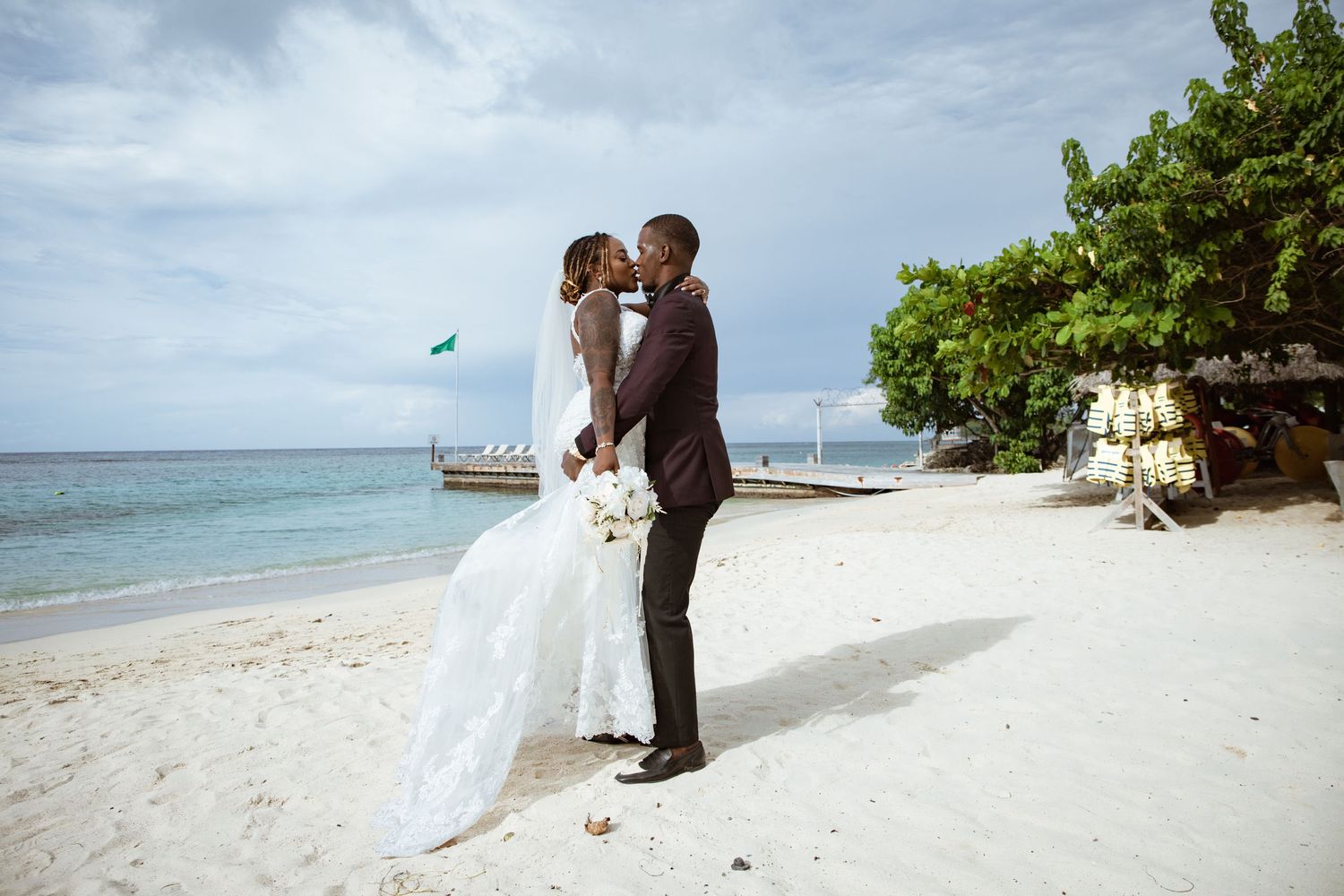Newlywed couple sharing a romantic kiss on a tropical beach with turquoise ocean waters in the background.