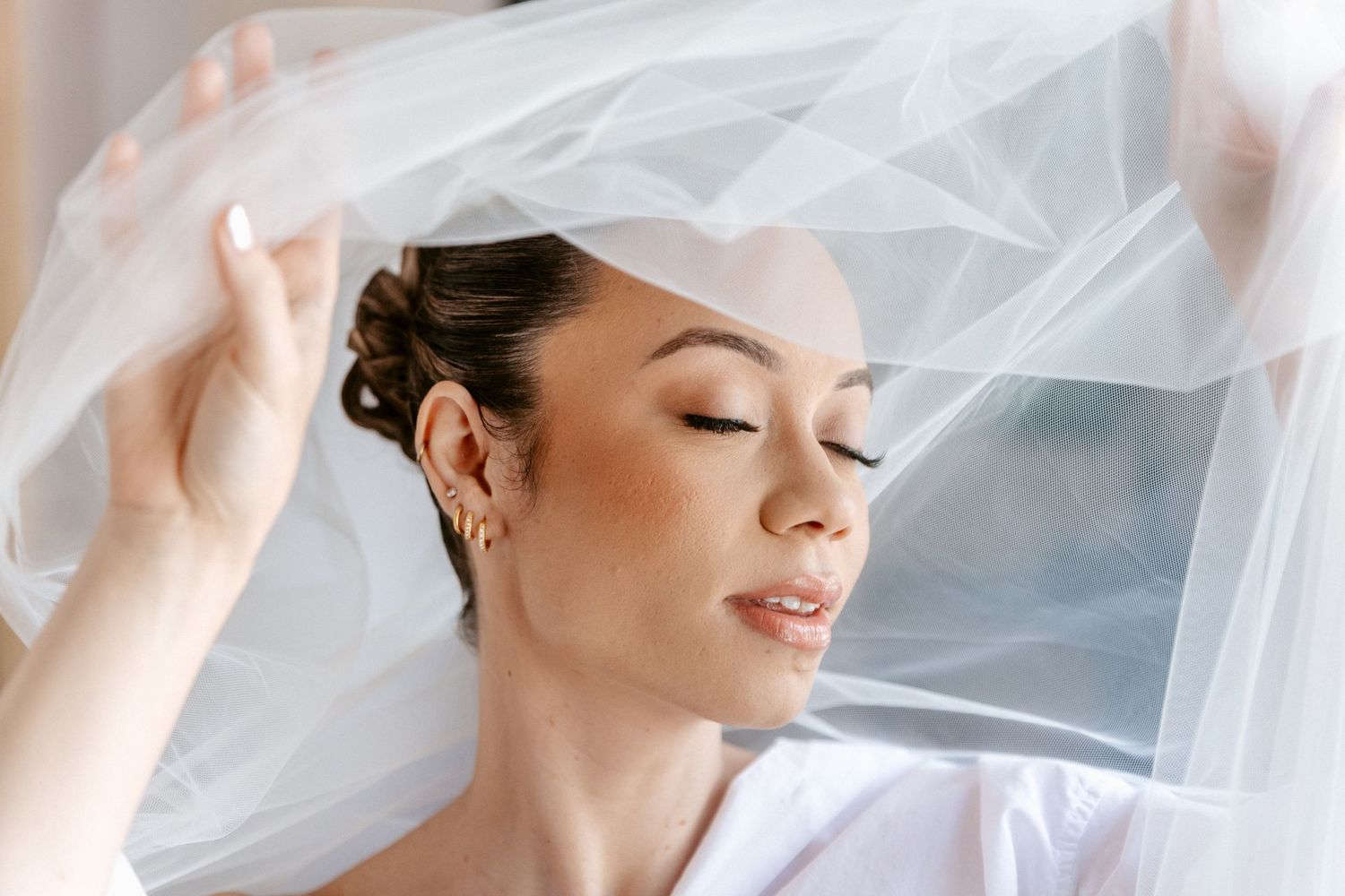 Bride with closed eyes lifting flowing white wedding veil in soft natural light.