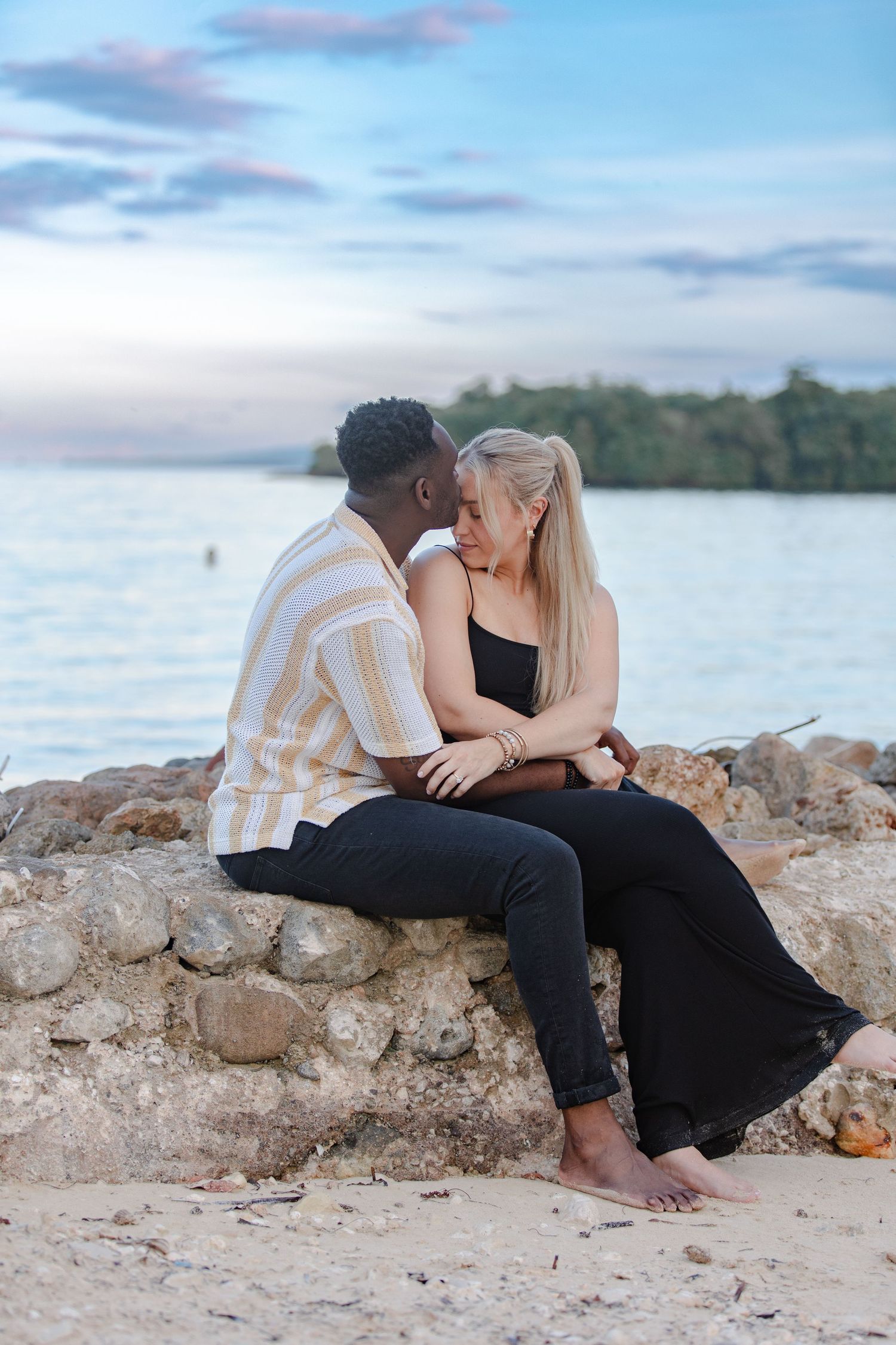 Romantic interracial couple sitting on a stone wall kissing near a tropical beach at sunset.