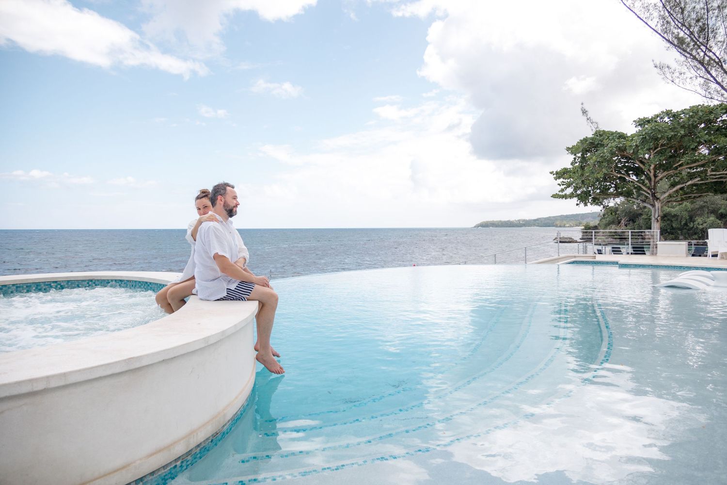 Couple sitting together on edge of infinity pool overlooking ocean at tropical resort.