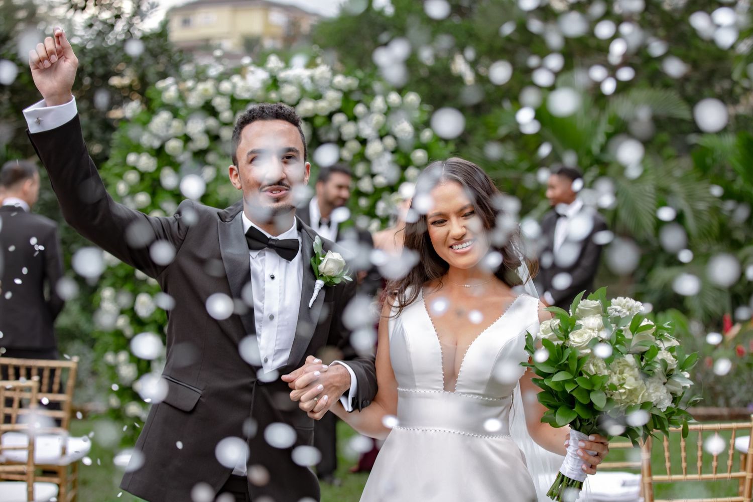 Joyful couple celebrates wedding exit with white confetti shower, bride holding bouquet outdoors.
