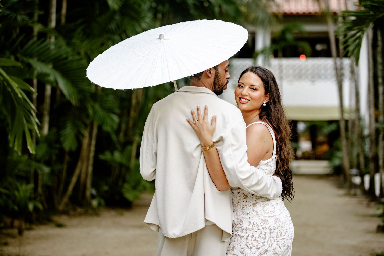 Smiling bride in white lace dress embraces groom holding white parasol umbrella outdoors among tropical greenery.