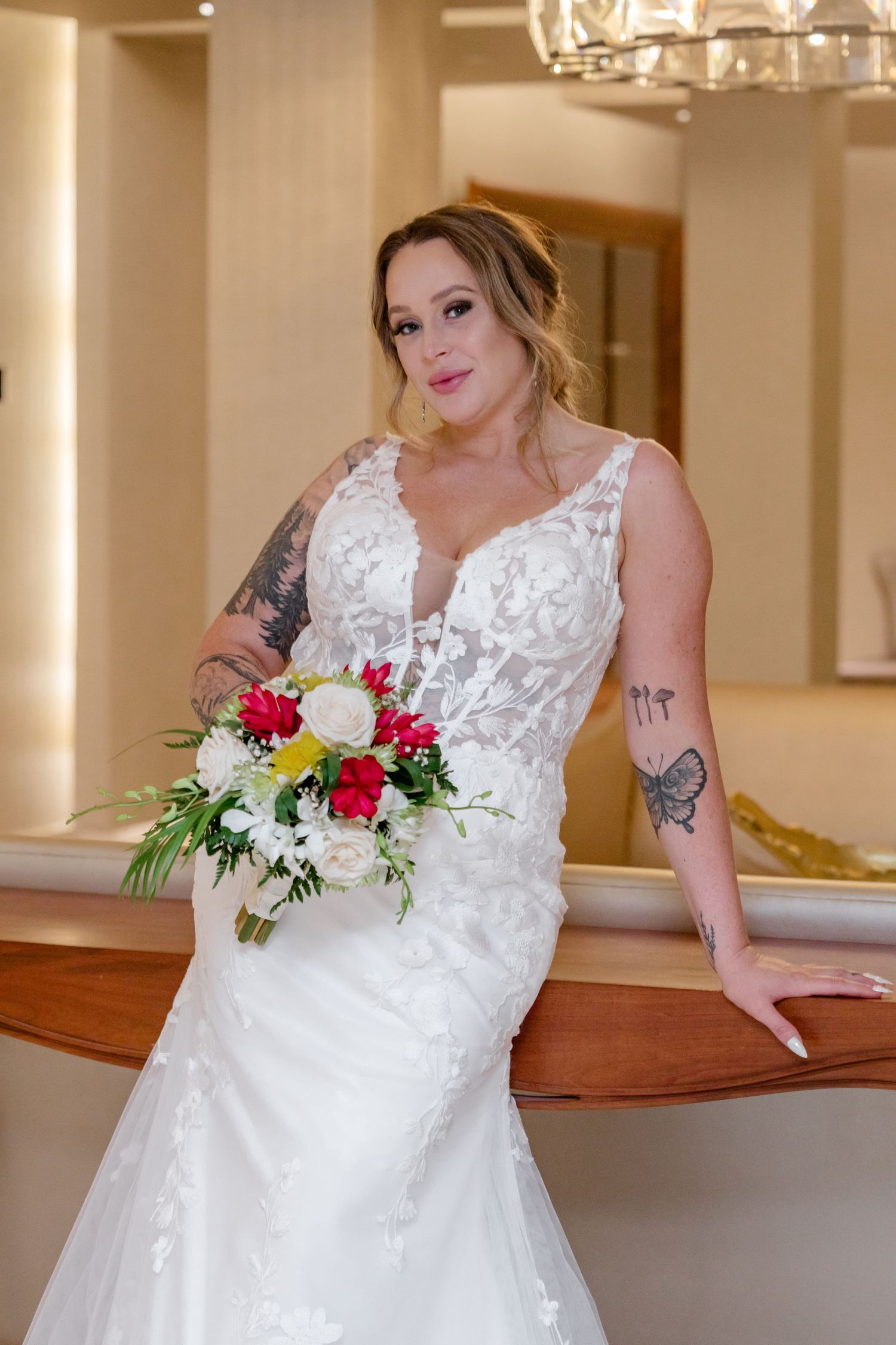 Bride in white lace mermaid gown holding colorful bouquet, tattooed arms, posing indoors at elegant wedding venue.