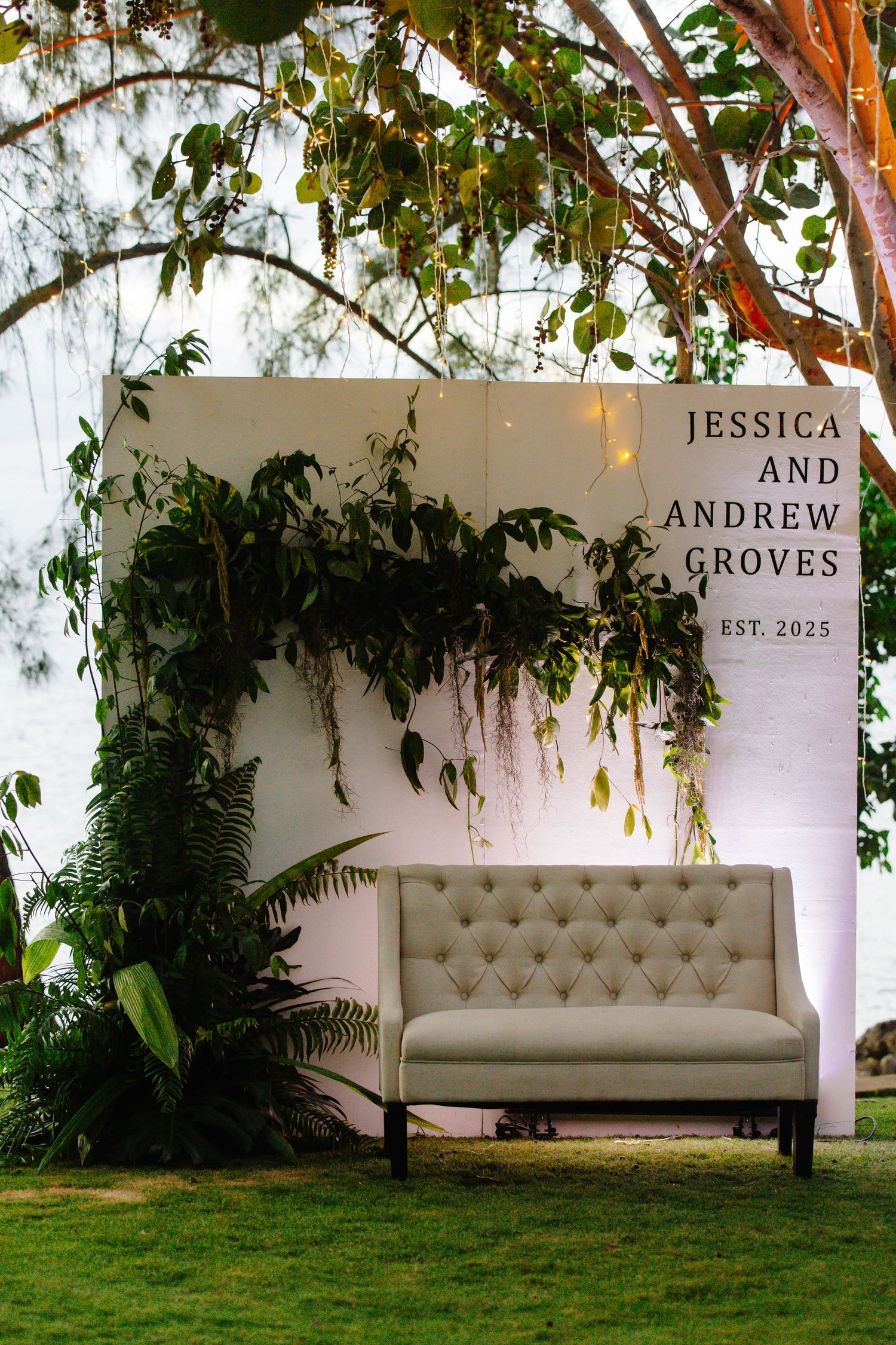 Elegant outdoor wedding photo booth with white backdrop, greenery, tufted loveseat, and Jessica and Andrew Groves Est. 2025 sign.